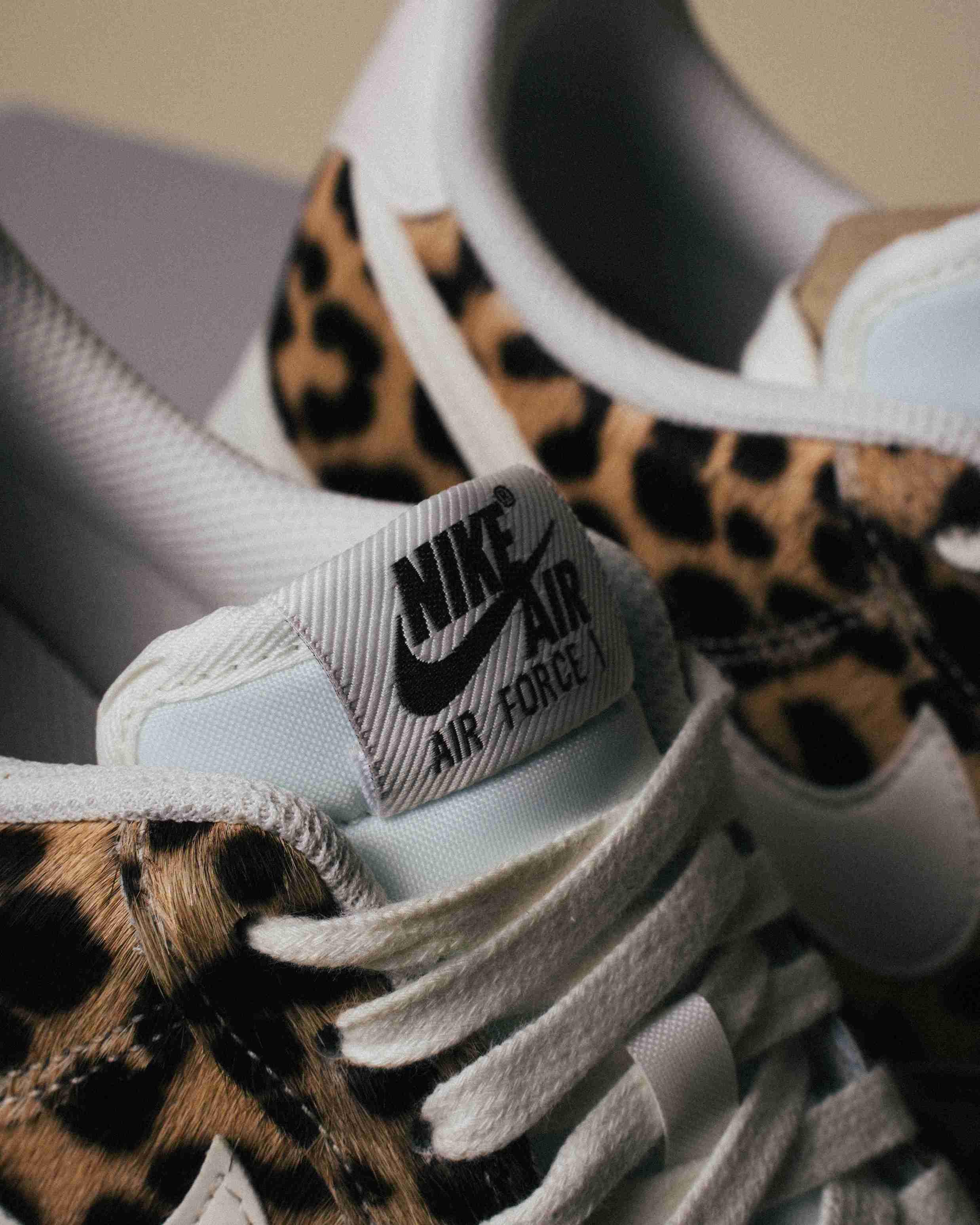 Paire de sneakers Nike Air Force 1 à motif léopard, placée à côté d’une boîte Nike grise sur un fond beige, vue artistique avec lumière douce.